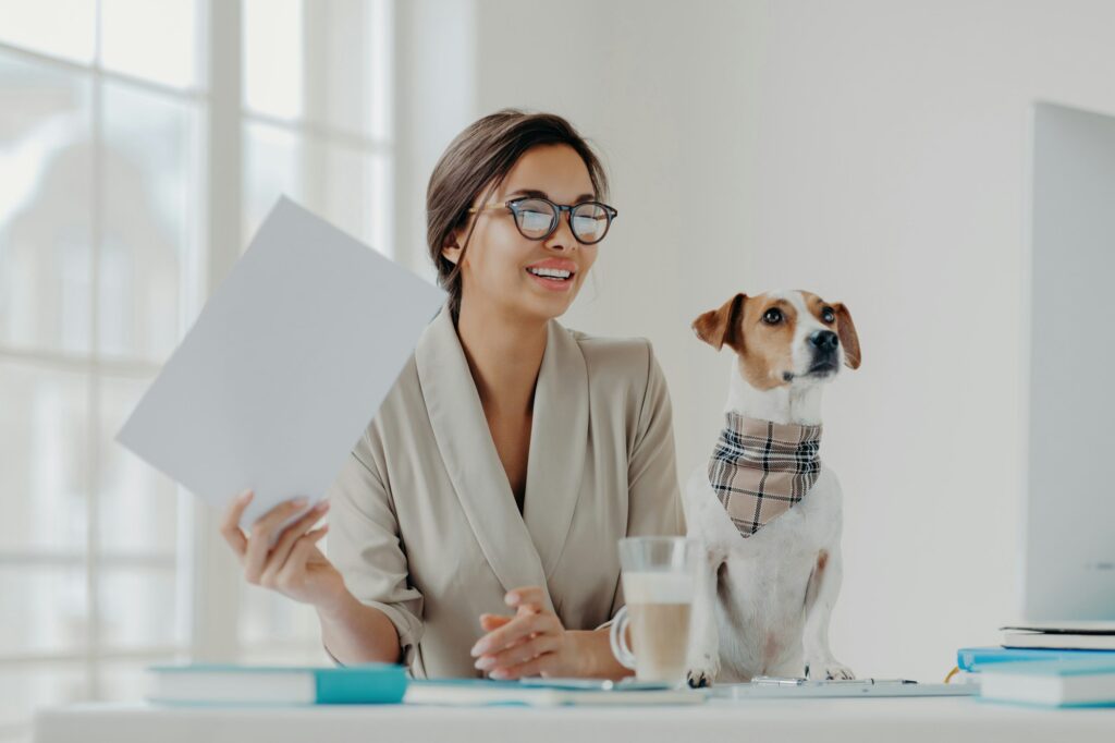 Smiling professional woman holding documents with her attentive dog at a bright home office